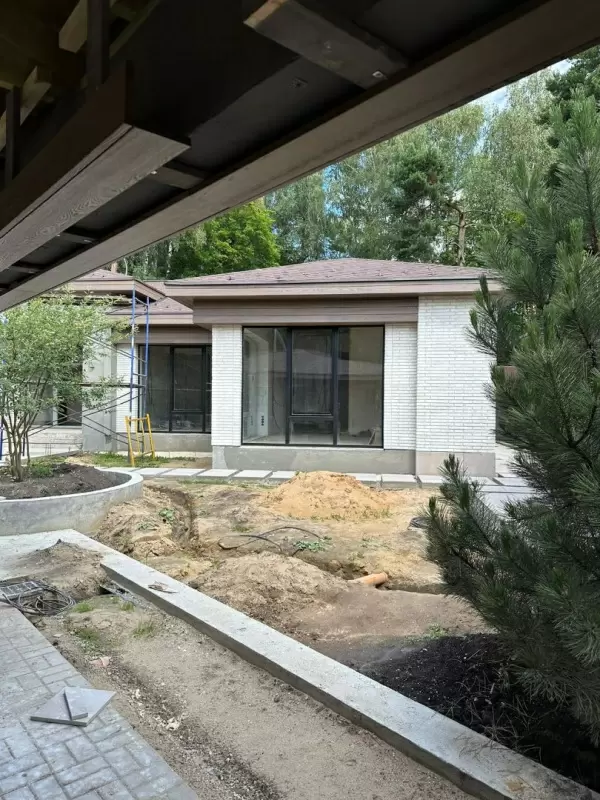 Отделка фасада частного дома с белой фасадной плиткой PETRA и большими окнами Процесс отделки фасада частного дома с использованием белой фасадной плитки PETRA, панорамных окон, окруженного деревьями и зеленью